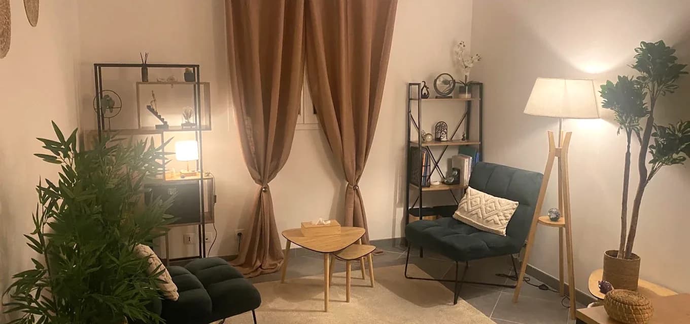Le cabinet de Cécile Gasparini à Célony : deux fauteuils en velours vert, une table basse en bois, une lampe allumée et des plantes.